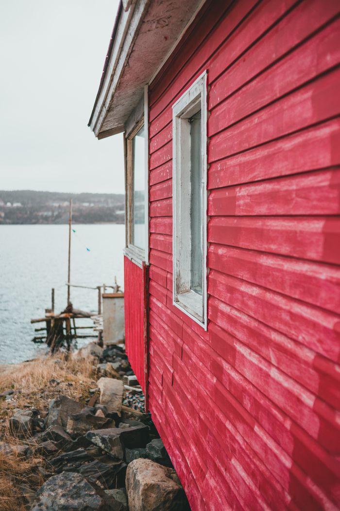 A rustic red wooden house stands by a calm riverbank, evoking a serene summer mood.